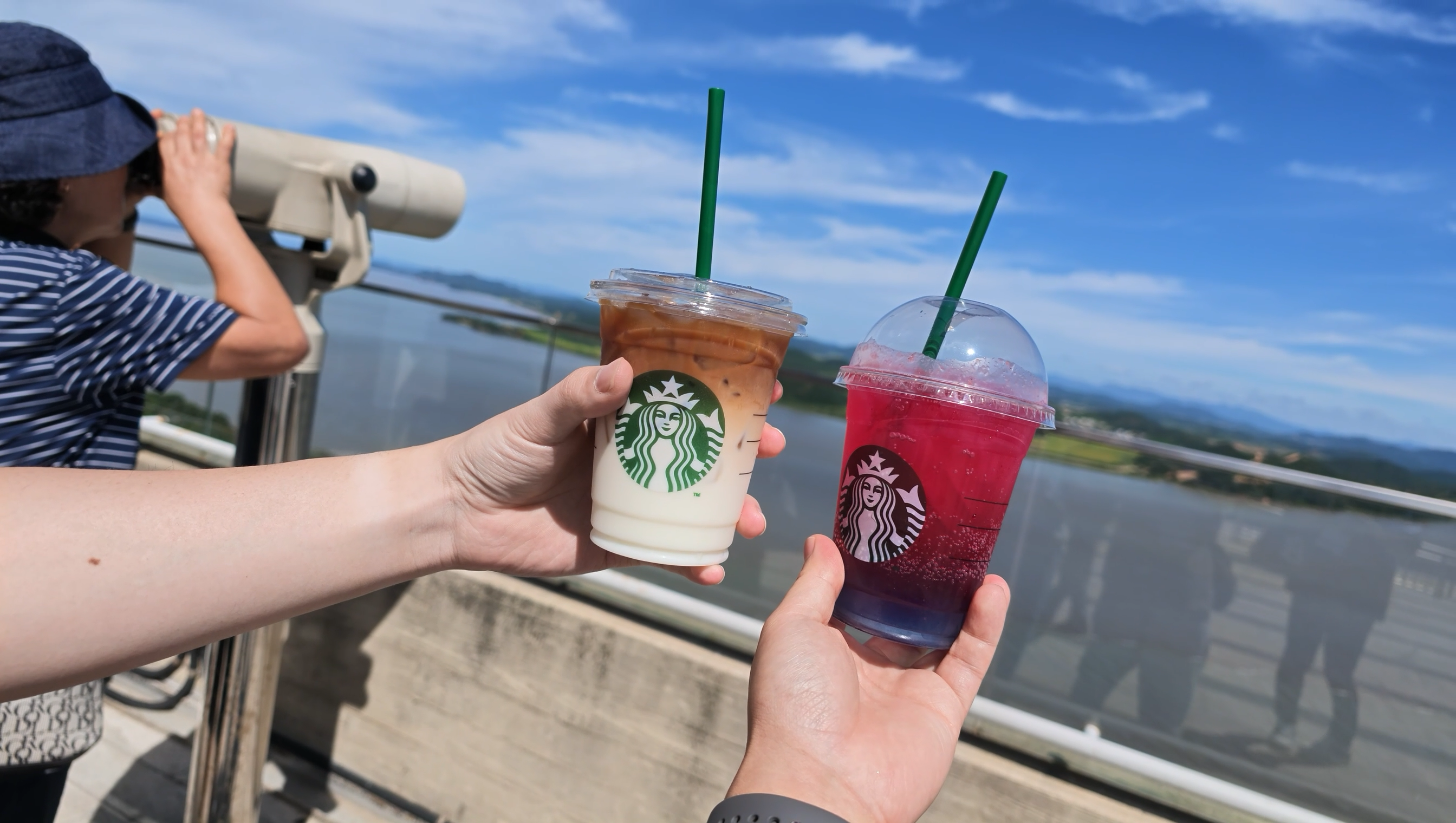 愛妓峰の展望台で、川と対岸の景色を背景にスターバックスのドリンク2杯を手に持った写真。横には望遠鏡をのぞく人がいる