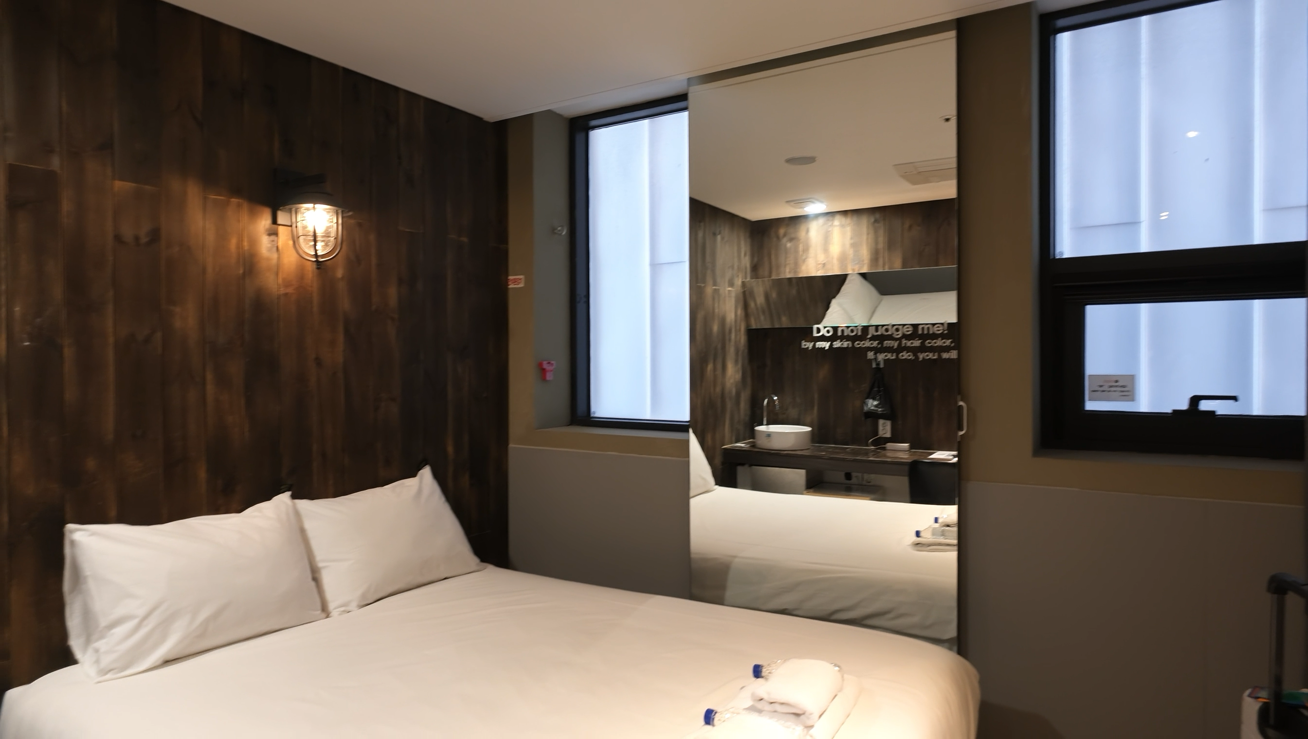 HERTZ Hotelの客室。木目調の壁に照明が付き、白いベッドの横には大きな鏡と窓がある