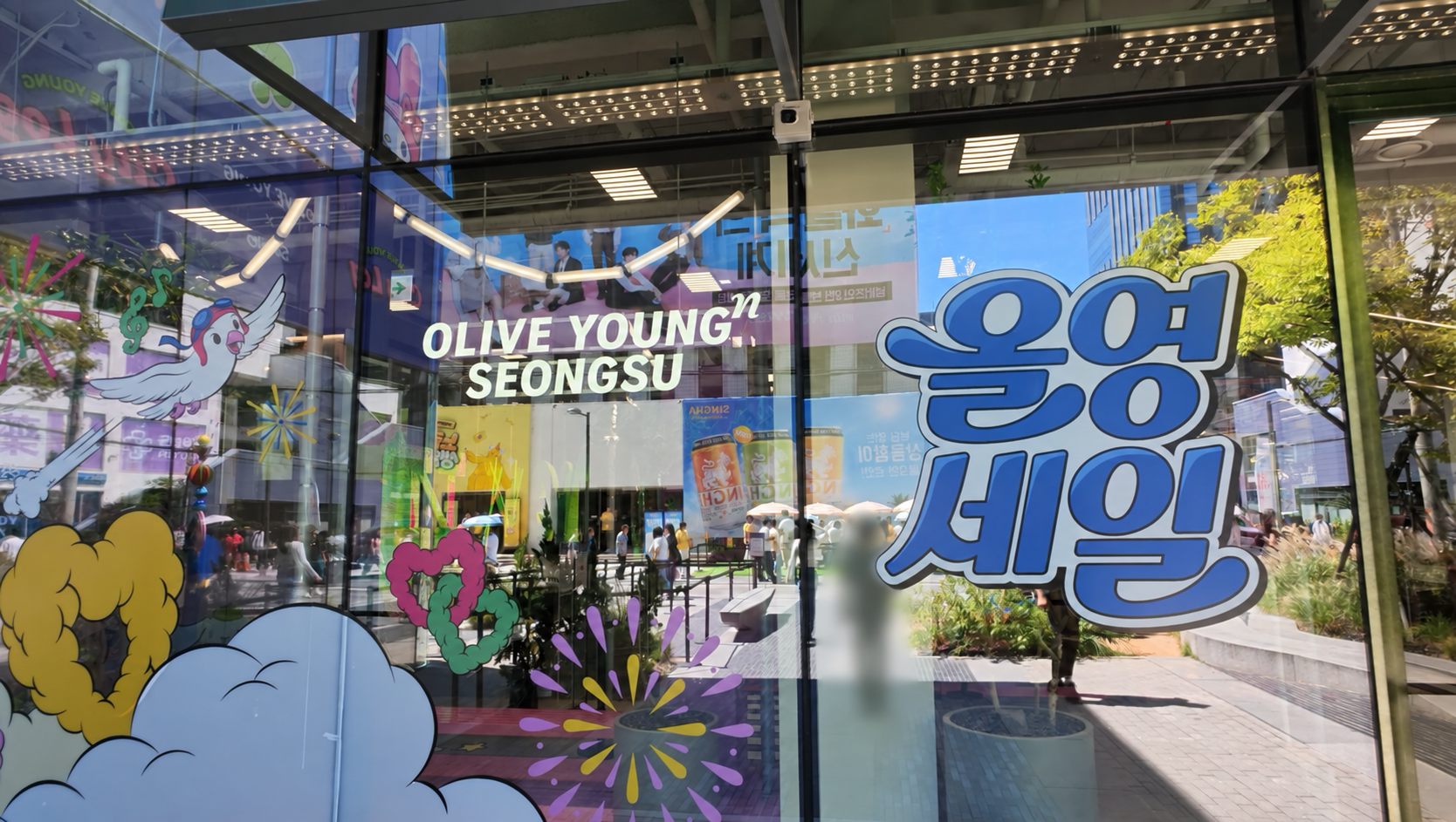オリーブヤングN聖水のガラス張りの店頭。OLIVE YOUNG N SEONGSUのロゴと、カラフルなイラストやセールの装飾が並んでいる