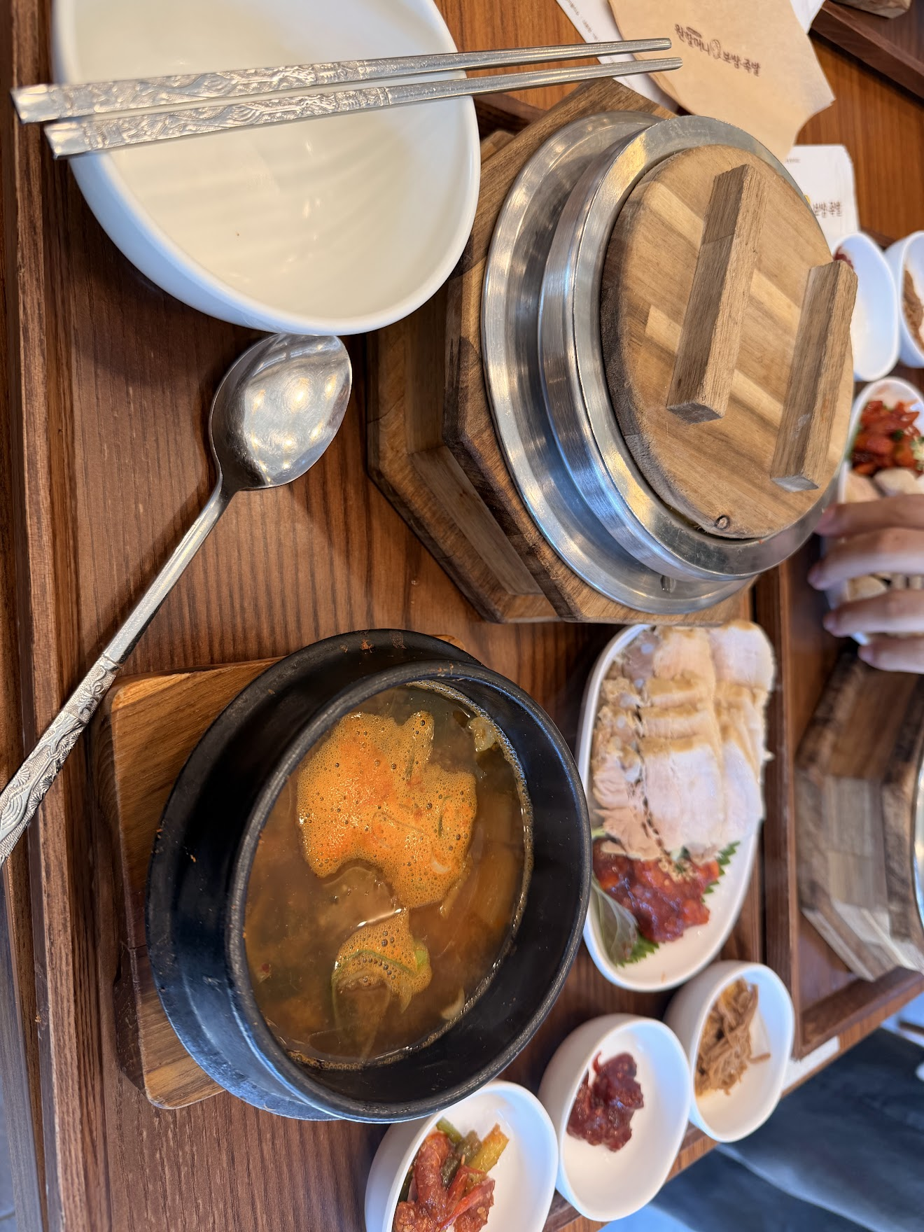 ポッサムの定食セットを上から撮影。薄切りの茹で豚が並んだポッサム、蓋付きの釜飯、オレンジ色の辛いスープ、そしてキムチやナムルなどのおかずが所狭しと並ぶ