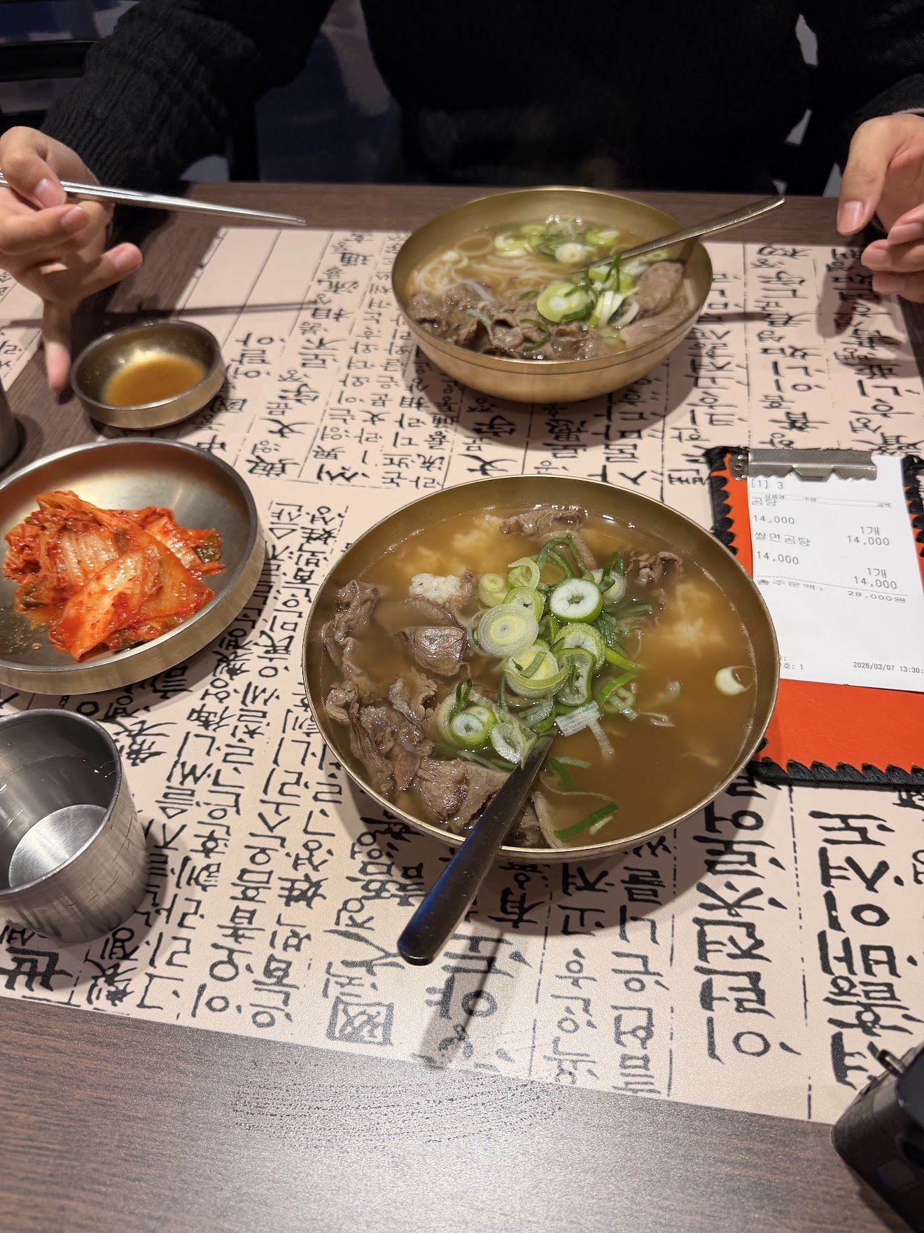 韓国語が書かれたテーブルマットの上に並ぶ2杯のコムタンスープ。牛肉とネギがたっぷり入った白濁のスープで、横にはキムチの小皿が添えられている
