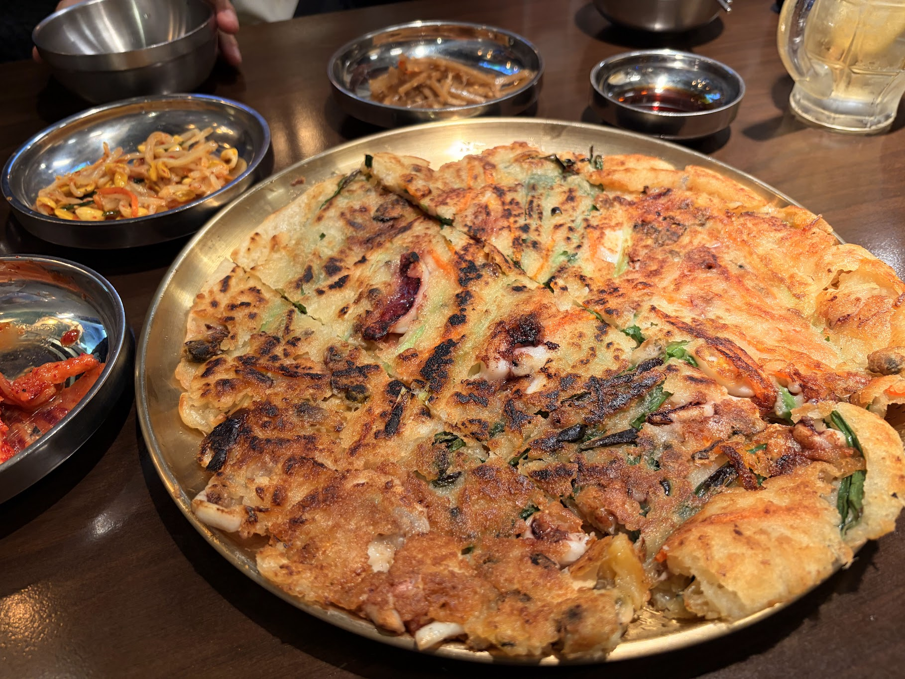 金色の大皿にどーんと盛られたこんがり焼けた海鮮チヂミ。周りにはキムチやナムルなどのおかずの小皿が並んでいる