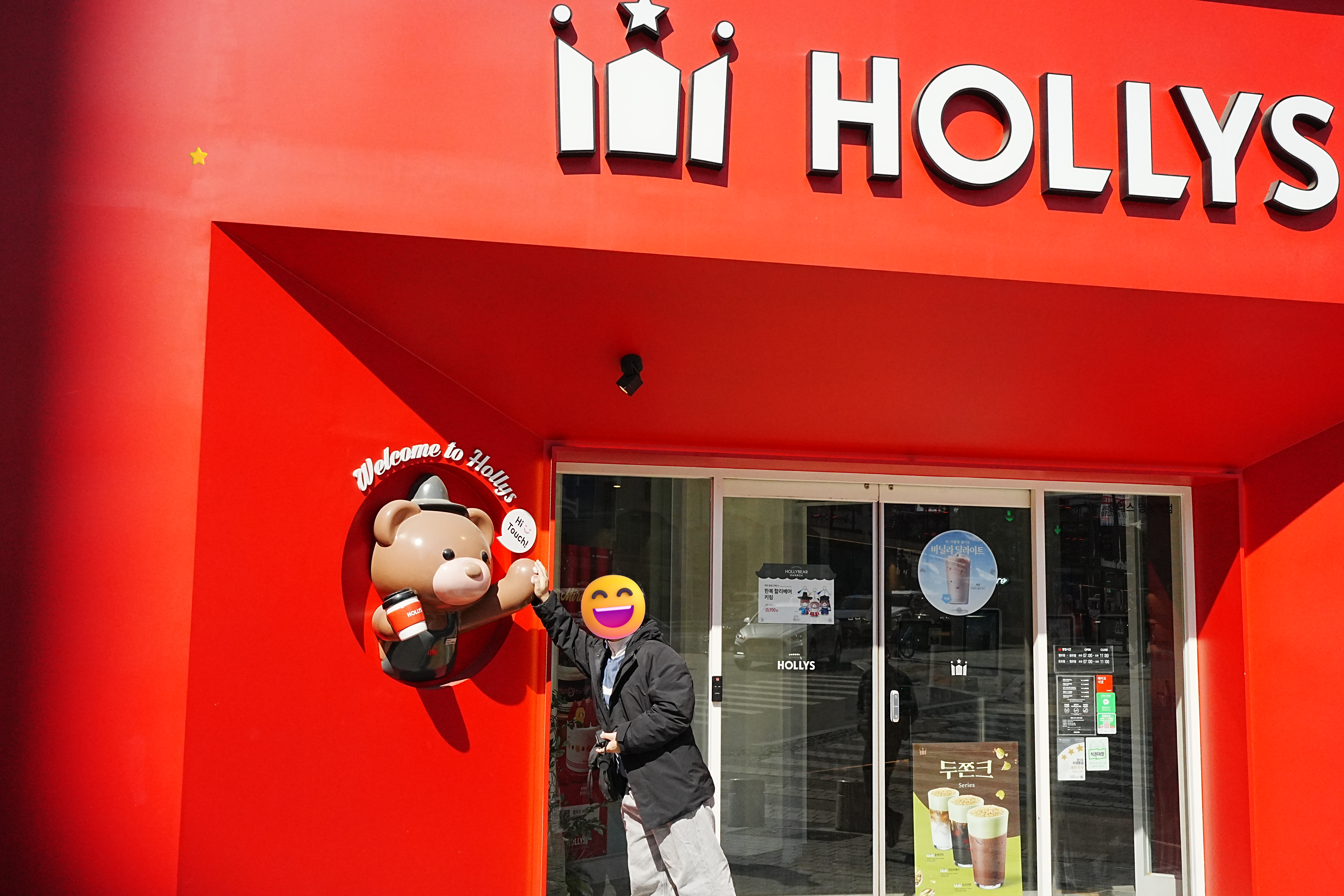 真っ赤な外観が目を引くカフェHOLLYSの入口前で、クマのマスコットキャラクターの横に立ってポーズをとる記念撮影
