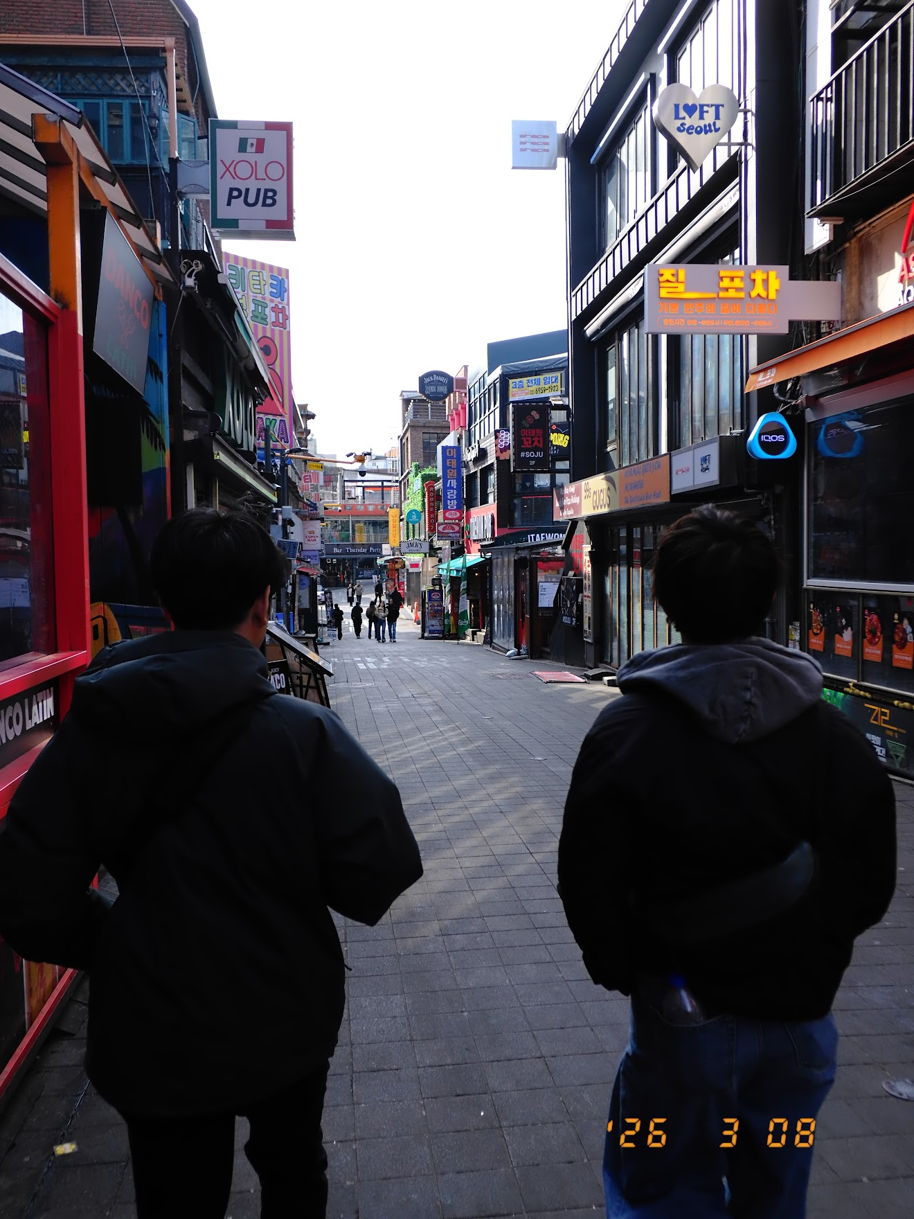 日曜日の昼間の梨泰院クラブ街を歩く友人二人の後ろ姿。看板やネオンが立ち並ぶ通りだが人はまばらで静か