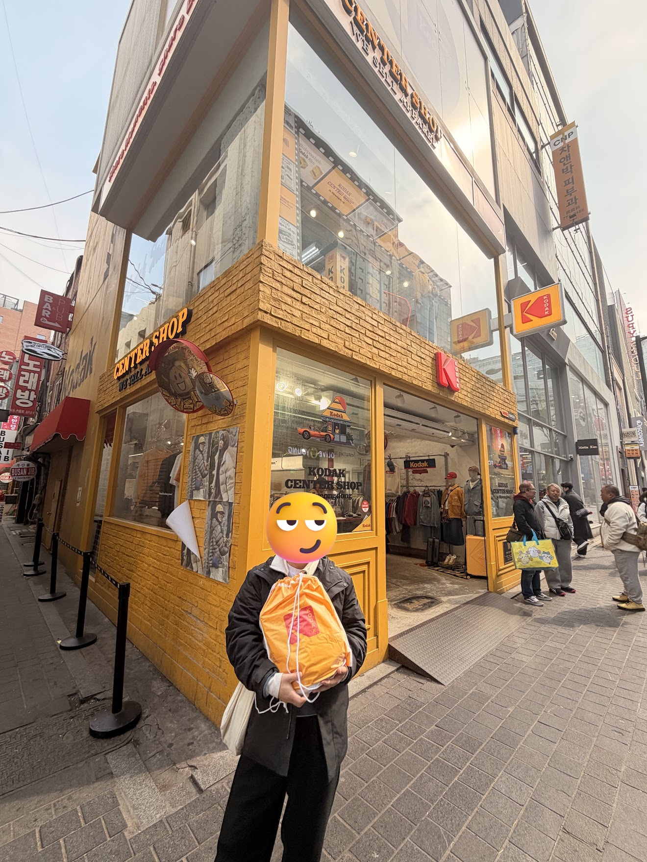 明洞のKODAK CENTER SHOPの黄色いレンガ調の店舗前で、オレンジ色の買い物袋を抱えて満足そうに立つ記念撮影。店頭にはKODAKのロゴが掲げられている
