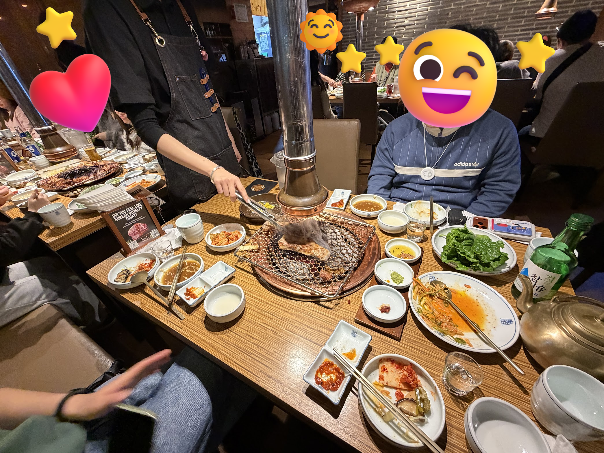 賑わう焼肉店で、黒いエプロンの店員さんがテーブル中央の炭火コンロでトングを使ってサムギョプサルを焼いてくれている。テーブルにはキムチやサンチュなどのおかずがぎっしり