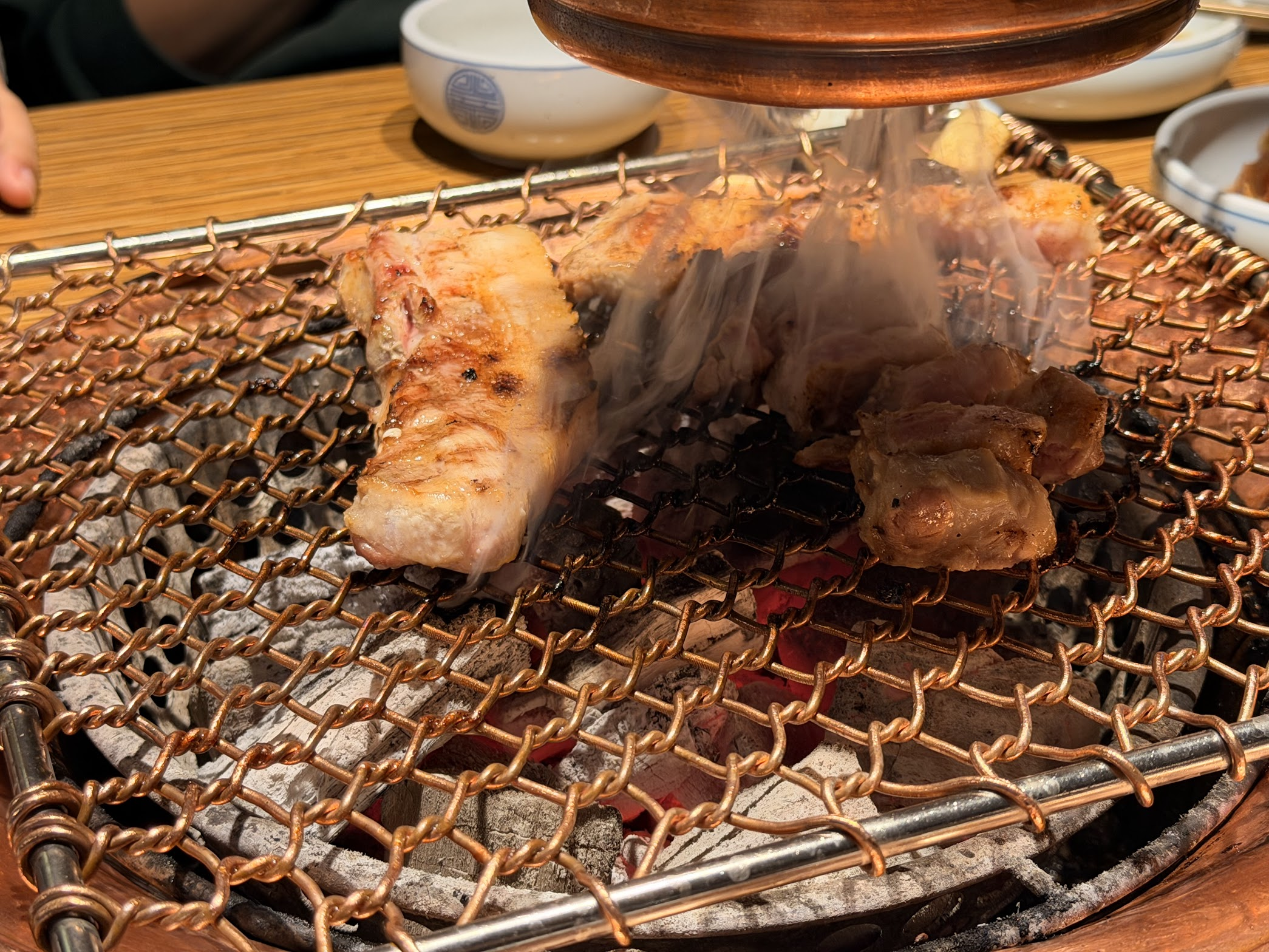 炭火の網の上で湯気を立てながらこんがり焼かれている厚切りサムギョプサルのアップ。炭の赤い火が網の隙間から覗いている